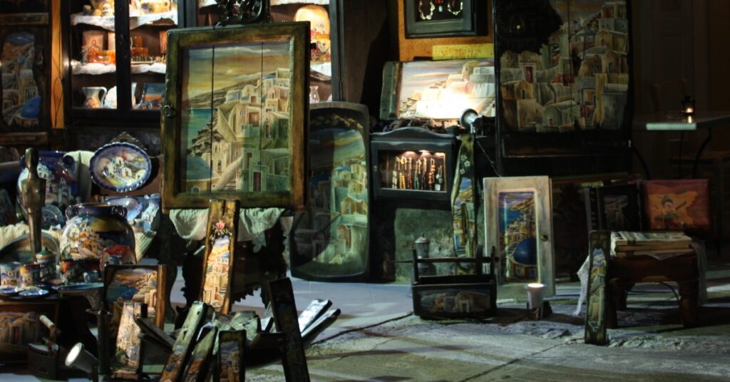photo of a gift shop in Oia, Greece. There are stacked up paintings and decorated plates on a crude dark wooden shelf. The rough hewn stone floor is visible - the atmosphere is evocative of an older time