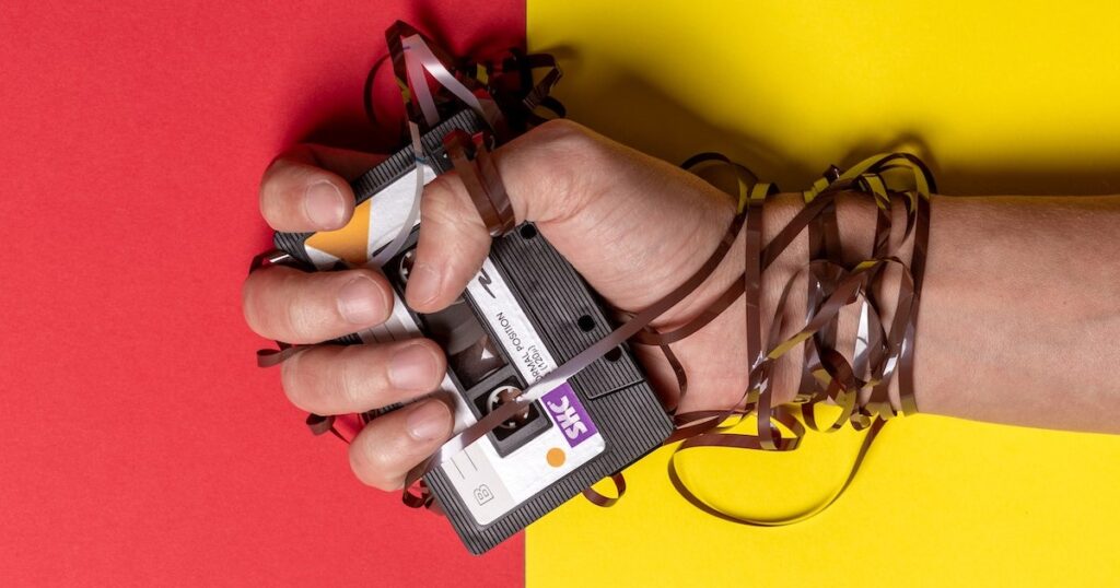 Hand holding a blank cassette tape, on a background of red and yellow block colours. Some of the magnetic tape is wrapped around the person's hands and wrists
