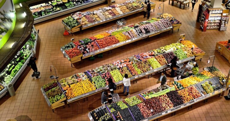 looking down at rows of supermarket tables packed with colourful fruits and veg. Spotlights have clearly been hung to pick out the food. A few people can be seen standing amongst the rows, looking at the options.