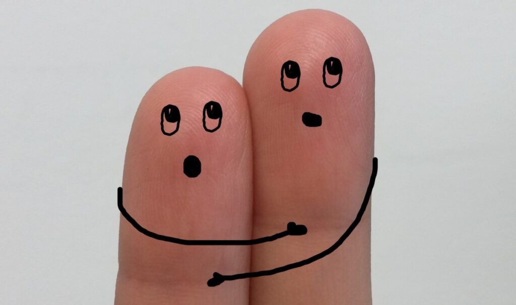 Closeup of two neighbouring fingertips from the same hand, resting against each other. Crude cartoonish eyes mouths and arms have been drawn on with a black pen, making it look like they are slightly surprised and hugging each other.