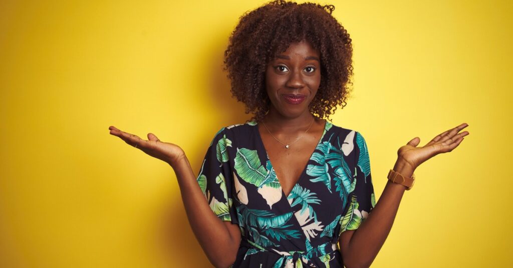 Woman making big 'shrug' gesture with a half-smile. She is wearing a dark dress with a print of leaves, and stands against a yellow background
