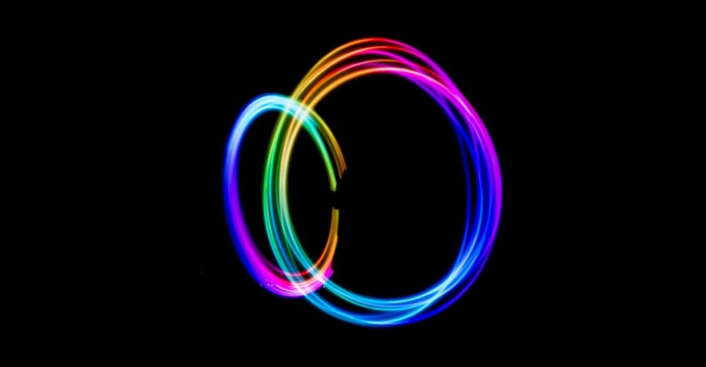 Two interlocking rings of multi-coloured glowing light (made with time-lapse photography of someone swinging lights that were changing colours in circles). various parts of the circles shift from yellow to pink to dark blue to light blue to cyan to green, and back to yellow