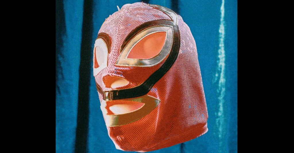 An empty red Mexican wrestler's mask floating against a background of a blue curtain. The fabric is slightly frayed, and it has gold rims around eliptical mouth and eye holes, and a black band that swoops around the top of the eyes, and down to meet across the top lip