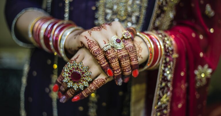 The hands and arms of a woman dressed ornately for a wedding in Pakistan. She has a red and purple robe covered in sequined flowers, she has multicoloured bangles on her arms, elaborate henna patterns drawn around her fingers, and four rings (from small to one that covers nearly her entire hand, that are encrusted with stones in circular and floral shapes