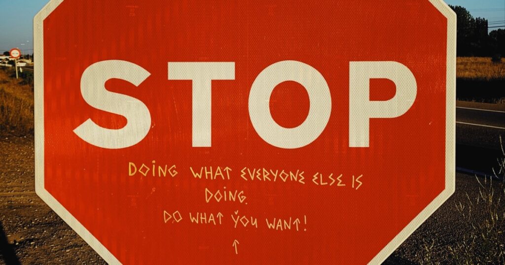 Closeup of a red octagonal STOP road sign. Underneath the word "STOP" someone has written with a white pen in block letters: "Doing what everyone else is doing. Do what you want!"