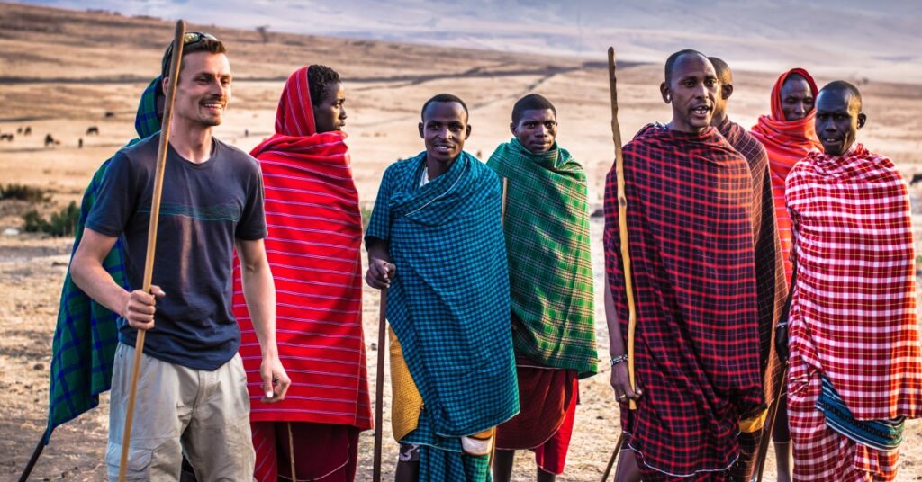 A smiling Caucasian man in t-shirt and khaki cargo shorts standing next to a group of 7 smiling (what appear to be) African Maasai tribesmen, wearing colourful body-wrap robes. Several of them are holding body-high walking sticks, including the man in the t-shirt.