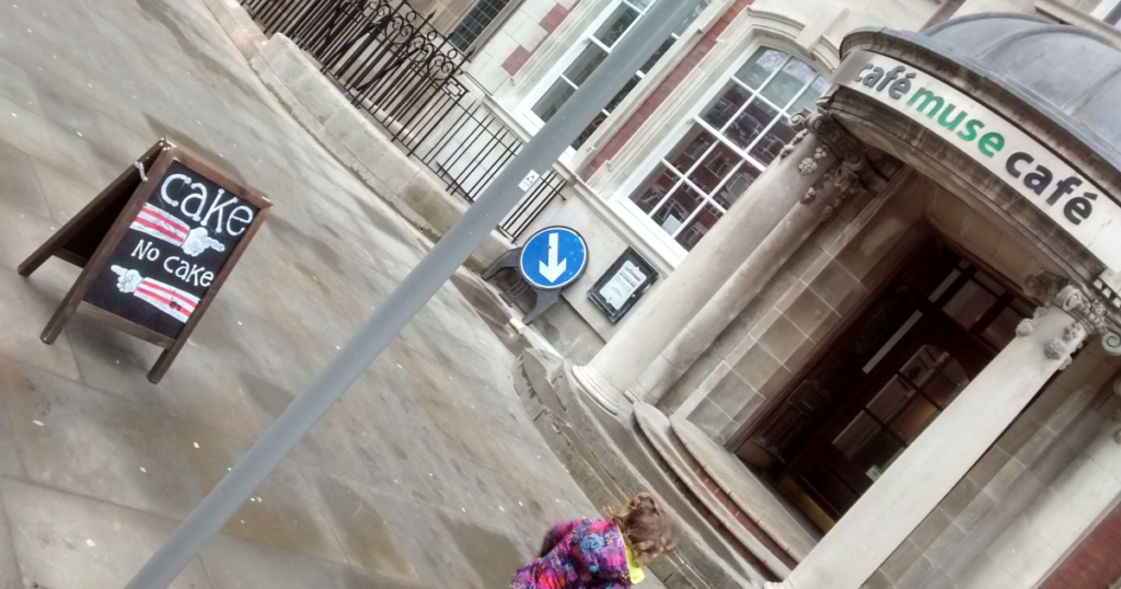 The outside of the Manchester Museum's cafe. A sign outside it has a hand drawn pointing at it which says "cake", and a hand pointing away saying "no cake"