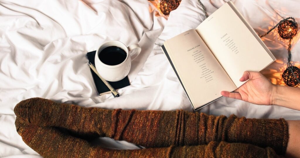 A woman sitting on bed reading poetry with a cup of coffee sitting on a diary with a pen resting on it. Only her hand and legs are visible, and she is wearing very cosy knee-length brown socks. Parts of a string of lights is seen lying on the bed too (cosy stylish ones, not colourful Christmas lights)