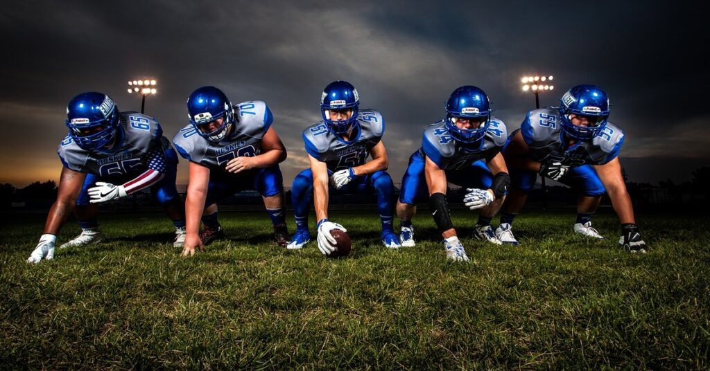 Five American football players in blue/grey uniforms squatting with their hands down as at a line of scrimmage, facing towards the camera as if they are about to run past it. The sky behind them is cloudy, and two floodlights are on