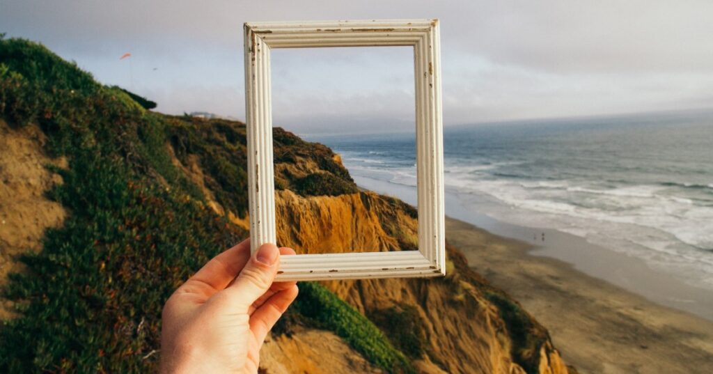 Landscape - cliffs over a sea. A hand is holding a white picture frame up in the middle of this, to isolate a pretty part of the cliff within its boundaries