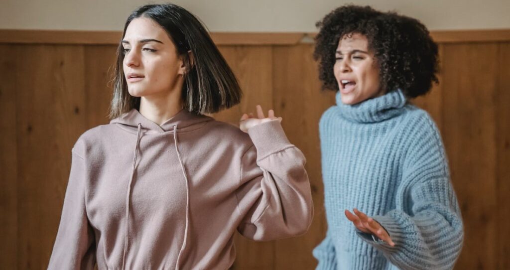 A woman in a blue sweater chases after a woman in a beige sweater, urgently trying to tell her something, The beige-sweater woman is turning her head away, and holding a hand to fend her off