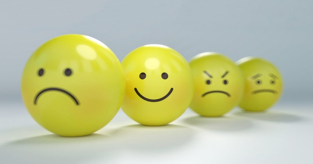 Four yellow balls against a plain grey background. Each has a different emoji face (sad, smiling, angry, scared). The smiley one is most in focus