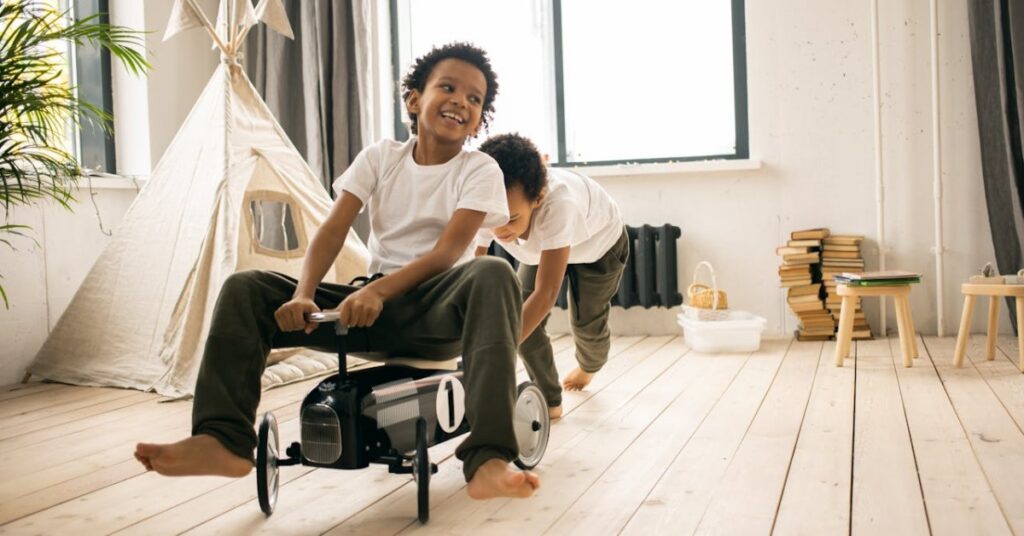Two boys playing in a light living room with a toy tent and stacked books in the background. One boy is sitting, smiling, on a toy car, steering it, while the other is behind the car and leaning forward almost into a crouch to push it quickly.