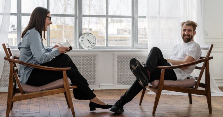 A man and a woman sitting in wooden deck chairs facing each other. They are inside on a hardwood floor, in front of a big bay window. The woman is wearing professional clothes and looking at an open notebook with a pen in her hand, and is laughing. The man is in a white t-shirt and dark trousers, and is looking directly at the camera with a bemused look. A plain white analogue clock sits on the windowsill behind them